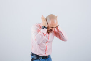 young man in pink shirt,jeans holding hands on ears while bending forward and looking crazy , front view.